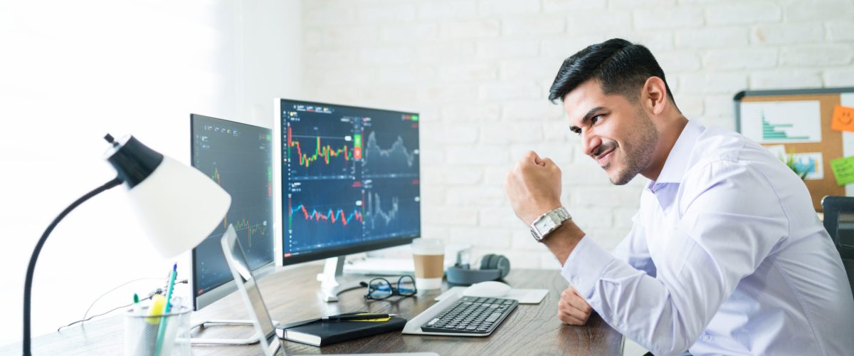 Confident handsome Hispanic financial broker showing fist to computer screen while trading from home